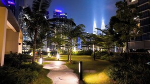 A nighttime view of a hotel courtyard with a pathway, palm trees, and modern buildings in the background. - JD Home Stay | Three Bedroom Apartment with Balcony @ Eaton Residence KLCC (Kuala Lumpur)