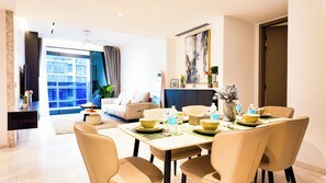 A modern dining area with a table set for four, a sofa in the background, and a view of the city through large windows. - JD Home Stay | Three Bedroom Apartment with Balcony @ Eaton Residence KLCC (Kuala Lumpur)