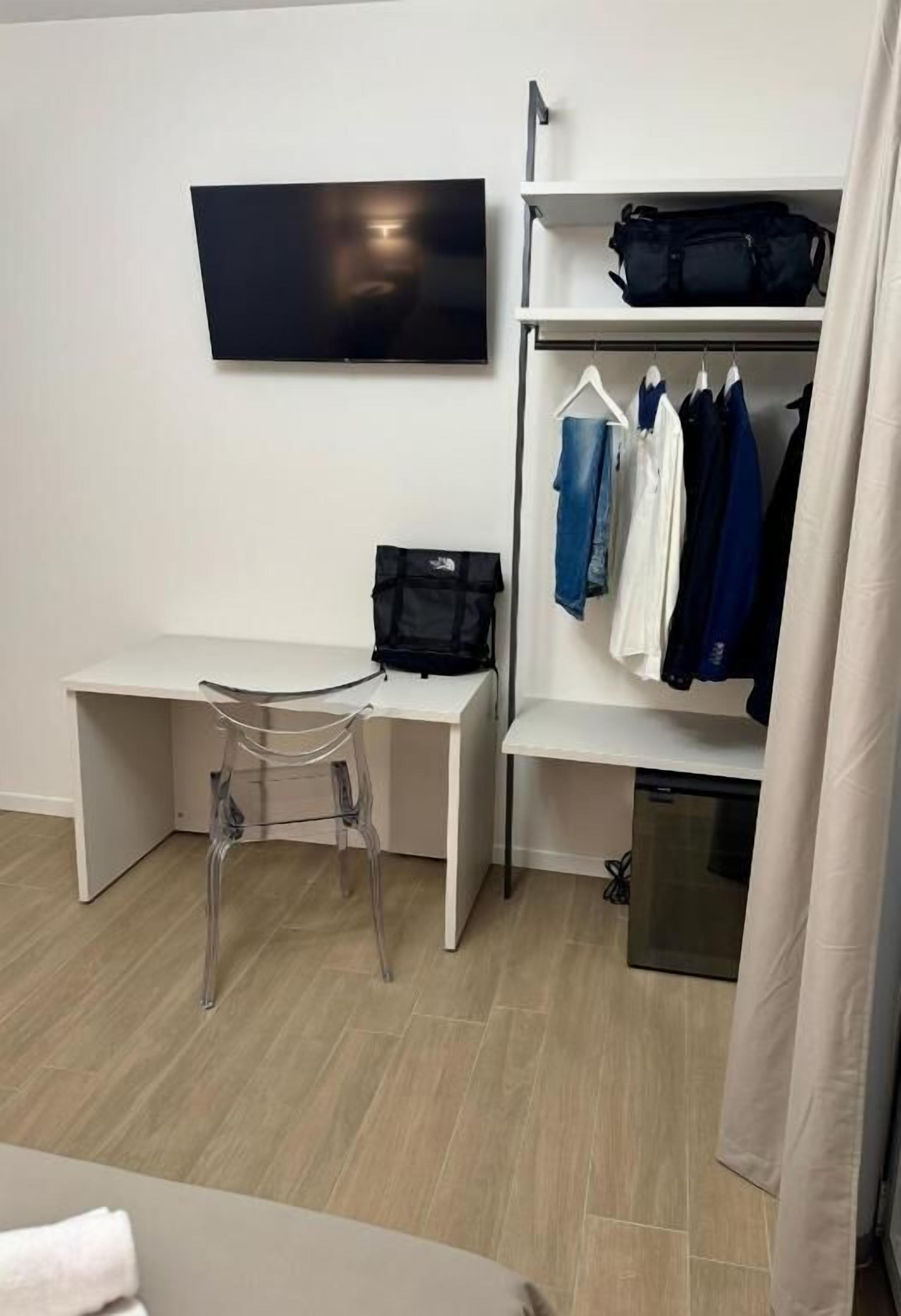 A hotel room with a desk, a chair, a TV mounted on the wall, a clothing rack, and a small cabinet.