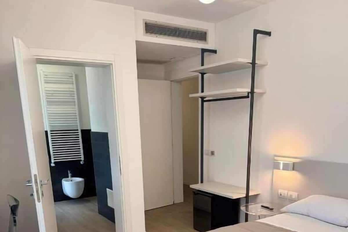 A hotel room with a bed, a bedside table, a shelf, a toilet, and a shower area.
