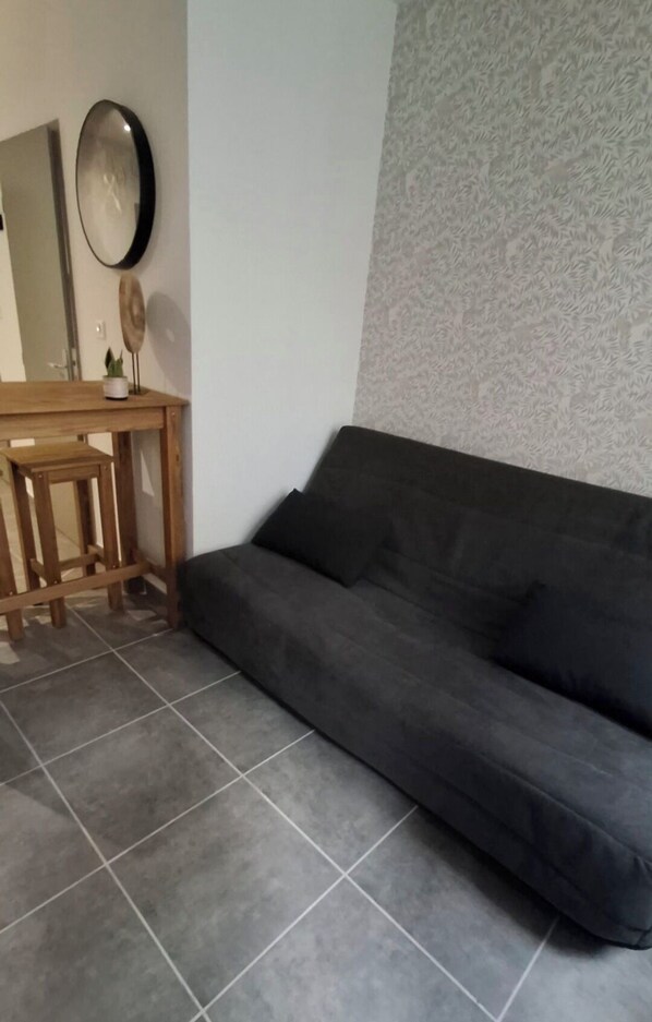 A room with a grey tiled floor, a dark grey sofa, a wooden table with a small plant, and a wall with a textured pattern.