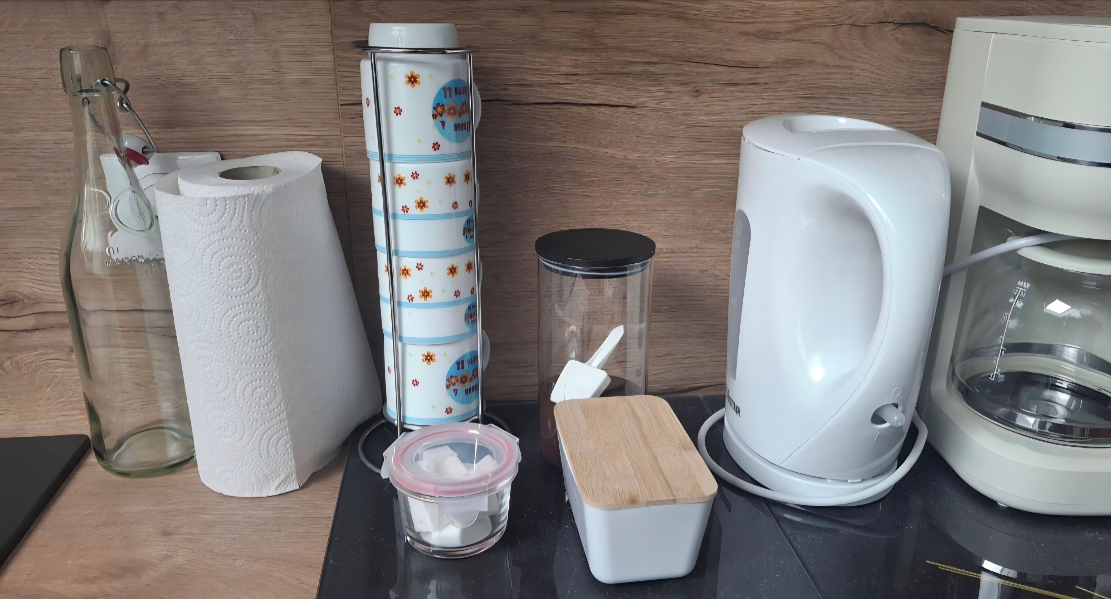 Una cuina amb un taulell, una cafetera, una ampolla de vidre, un rotllo de paper absorbent, un recipient cilíndric amb tapa i una petita caixa de fusta.