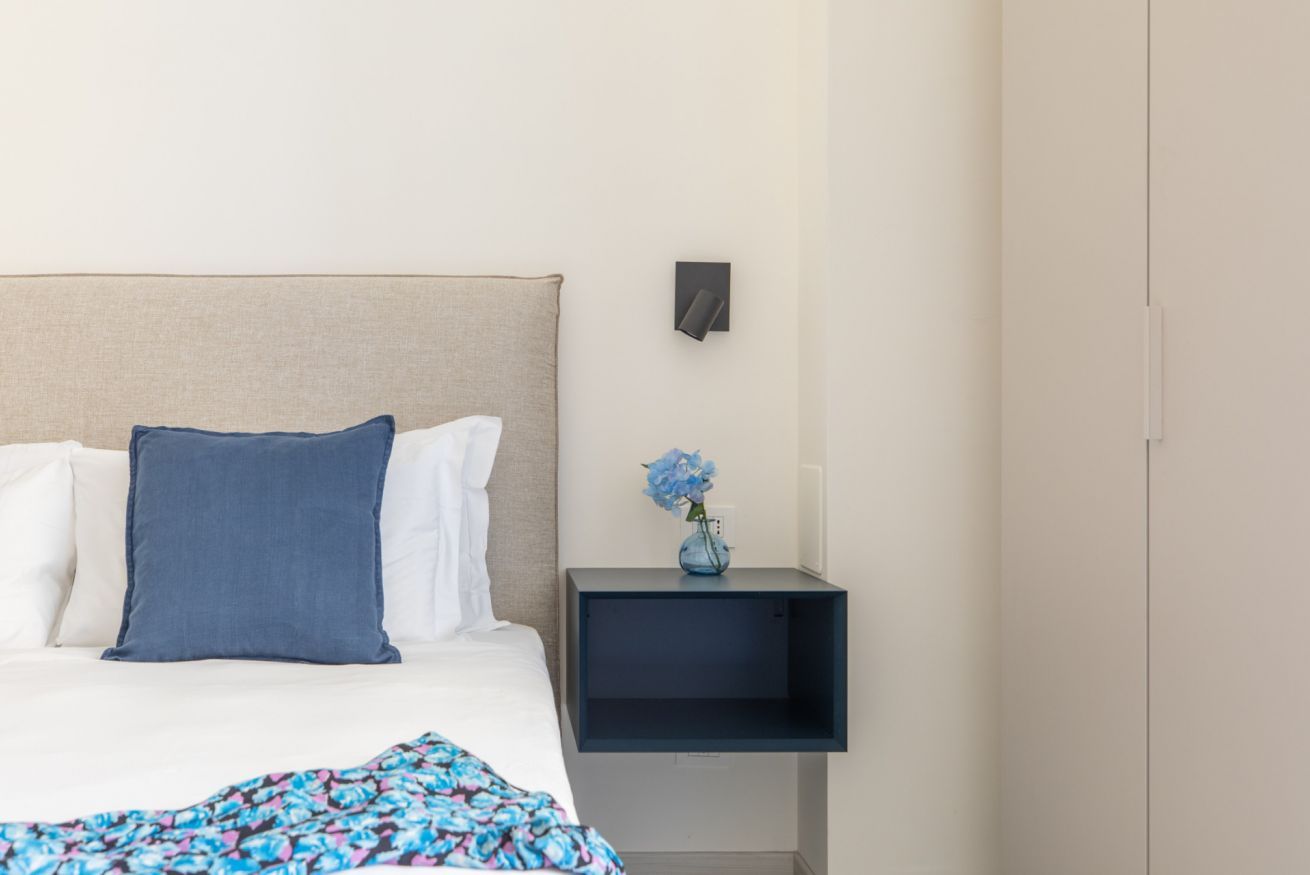 A neatly made bed with white and blue bedding, a patterned throw, a small bedside table with a vase of flowers, and a wall-mounted light fixture.