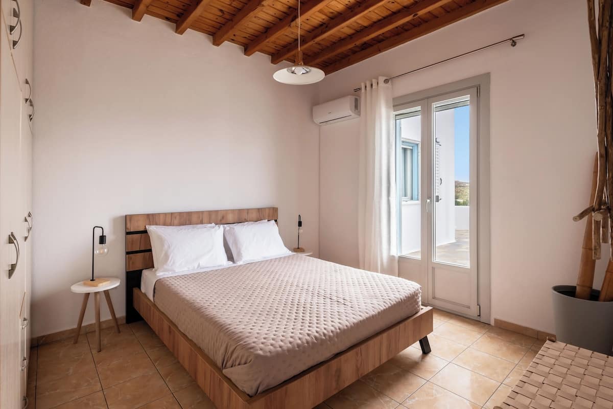 Quarto com cama de madeira, mesa de cabeceira pequena, ar condicionado fixado na parede e vista para o exterior por uma porta deslizante.