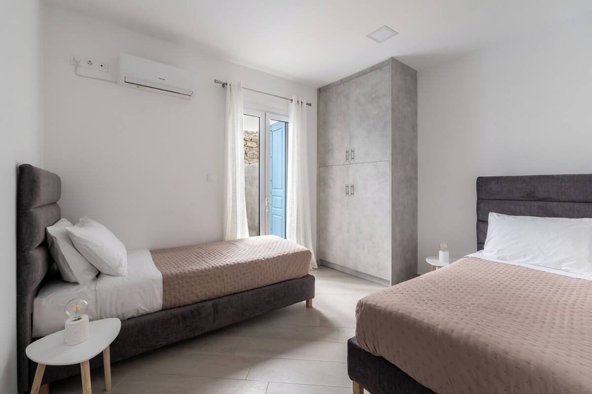 Quarto de hotel moderno com duas camas, um guarda-roupa, uma mesa pequena e uma janela com cortinas.