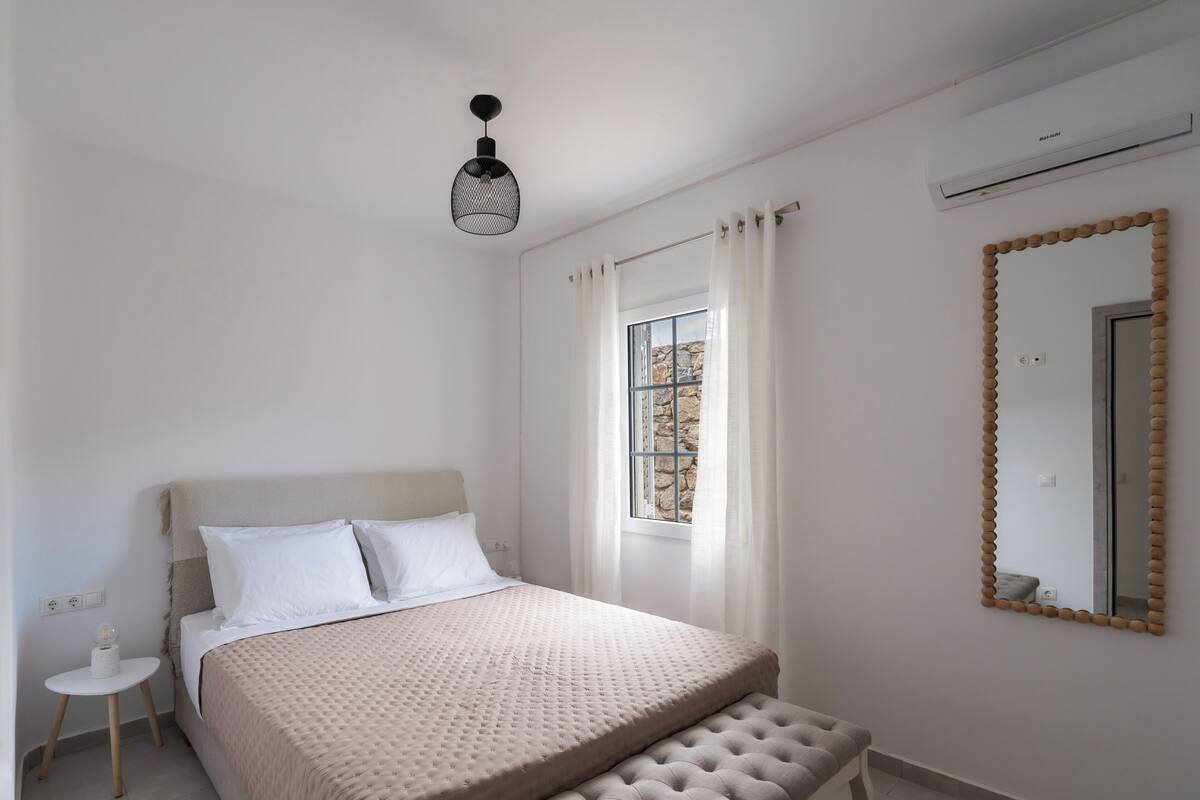 Quarto com cama, mesa de cabeceira, banqueta, janela com cortinas e aparelho de ar condicionado.