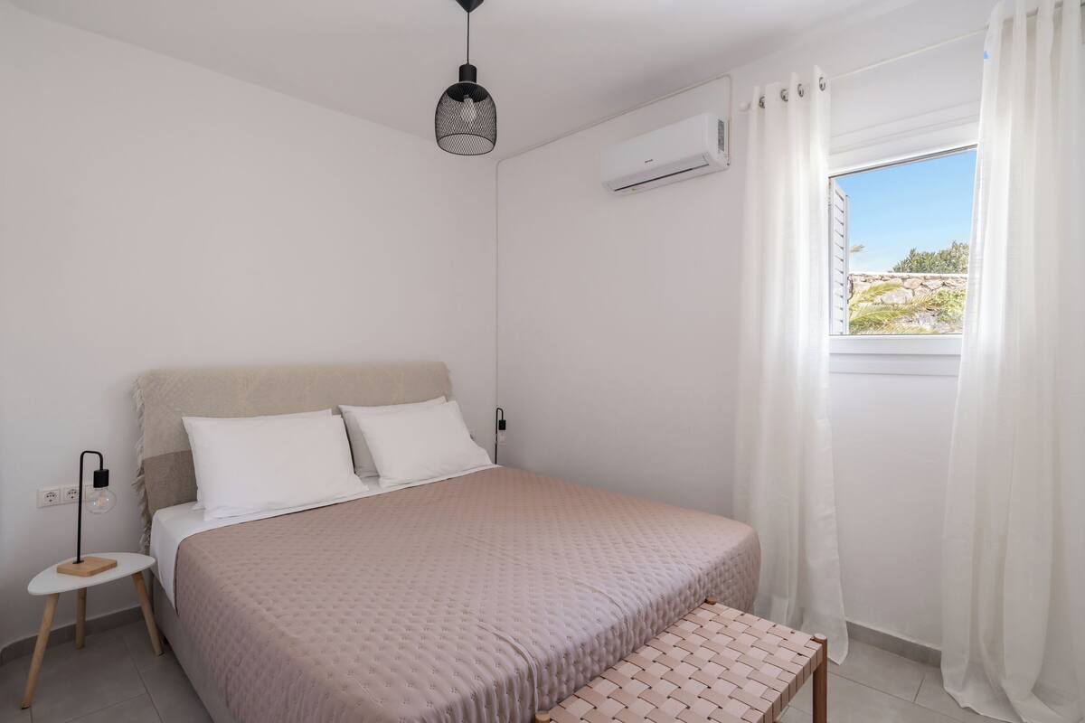 Quarto com cama, mesa de cabeceira, aparelho de ar condicionado e janela com cortinas.