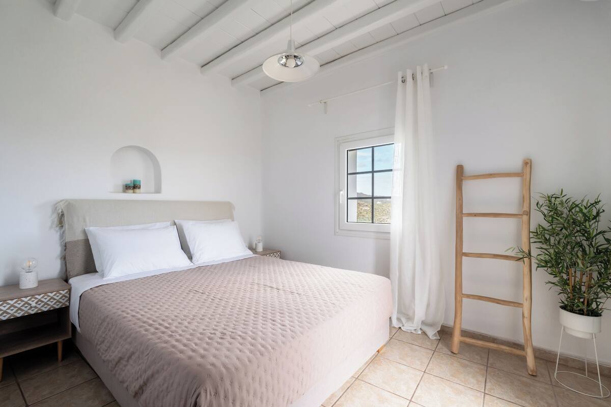 Quarto com cama, mesa de cabeceira, janela com cortinas, escada de madeira e um vaso com planta.