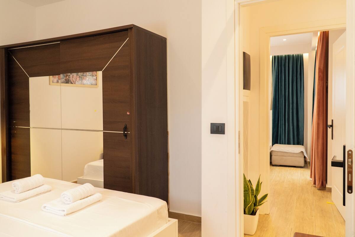 A hotel room with a bed, a wardrobe, a mirror, a potted plant, and a view into another room.