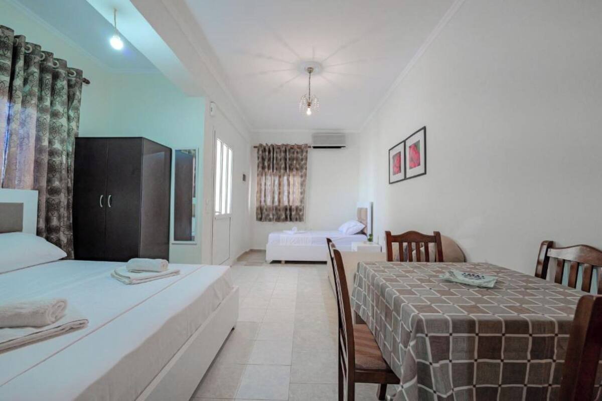 A bedroom with a bed, a wardrobe, a dining table with chairs, and a small kitchenette area.