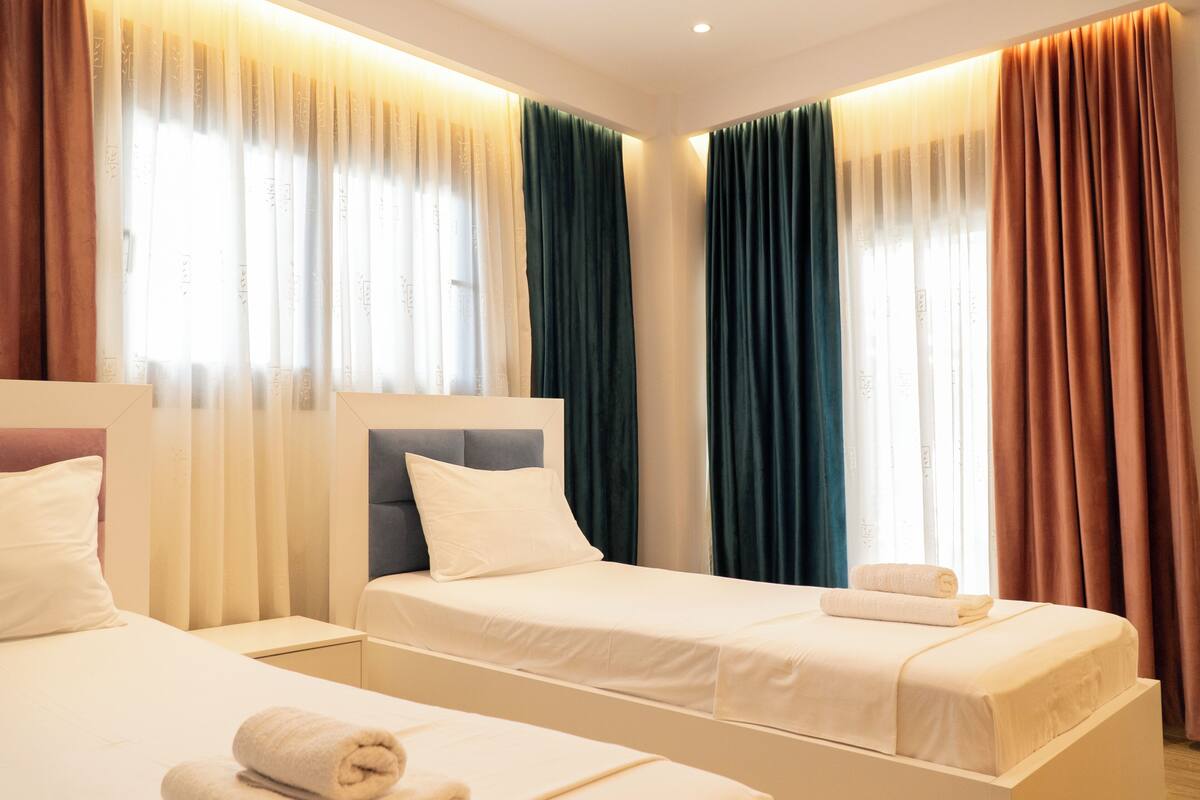 A hotel room with two single beds, a headboard, a nightstand, and a window with curtains.