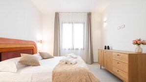 A neatly made bed with a beige bedspread, a wooden dresser with drawers, and a window with curtains. - 'Luxi Teulada' Apartment with Wi-Fi and Air Conditioning (Teulada)