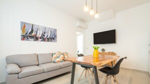 A modern living room with a sofa, a wooden dining table, and a flat-screen TV mounted on the wall. - 'Luxi Teulada' Apartment with Wi-Fi and Air Conditioning (Teulada)