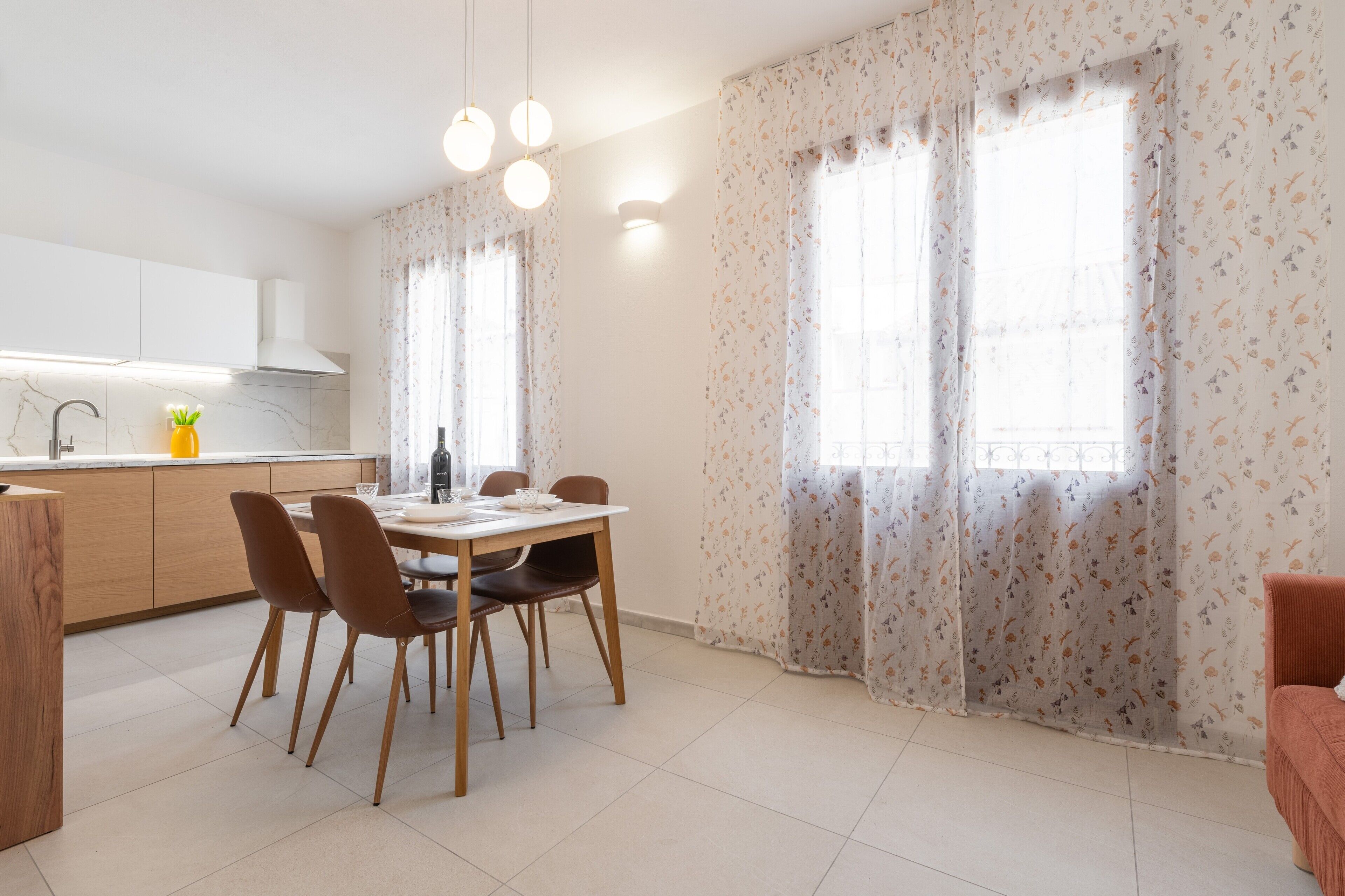 Une cuisine moderne avec des meubles blancs, une table à manger avec des chaises et une fenêtre ornée de rideaux à motifs floraux.