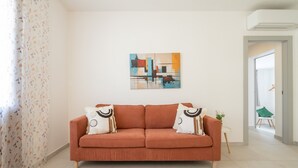 Una sala de estar con un sofá, un cuadro en la pared, una planta y vistas a otra habitación.