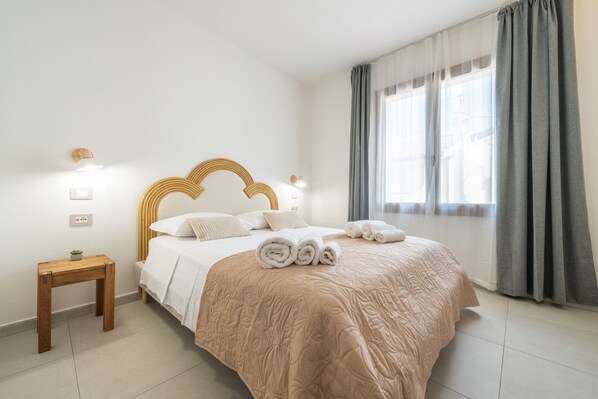 A neatly made bed with a headboard, a bedside table with a plant, and a window with curtains. - The Essence of Light in a Designer Apartment in Teulada (Teulada)
