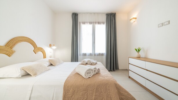 Una habitación de hotel con cama, mesitas de noche, cómoda, ventana con cortinas y una lámpara fijada a la pared.