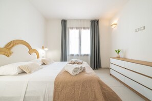 A hotel room with a bed, bedside tables, a dresser, a window with curtains, and a wall-mounted light fixture. - The Essence of Light in a Designer Apartment in Teulada (Teulada)