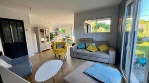 Una sala de estar moderna con sofá, sillón y reposapiés, un comedor con sillas y una cocina al fondo.