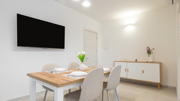 Uma área de refeições com uma mesa de madeira, cadeiras brancas, uma TV na parede, um armário branco e um vaso com flores.