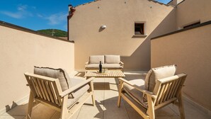 Um pátio externo com móveis de madeira, uma mesa e um edifício bege ao fundo.