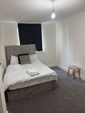 A neatly made bed with a grey headboard, a small wooden stool, and a window with dark curtains. - Central Luxurious Apartment London Bridge (London)