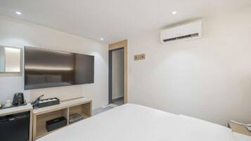 A hotel room with a bed, a TV mounted on the wall, a small desk with a coffee maker, and an air conditioning unit.