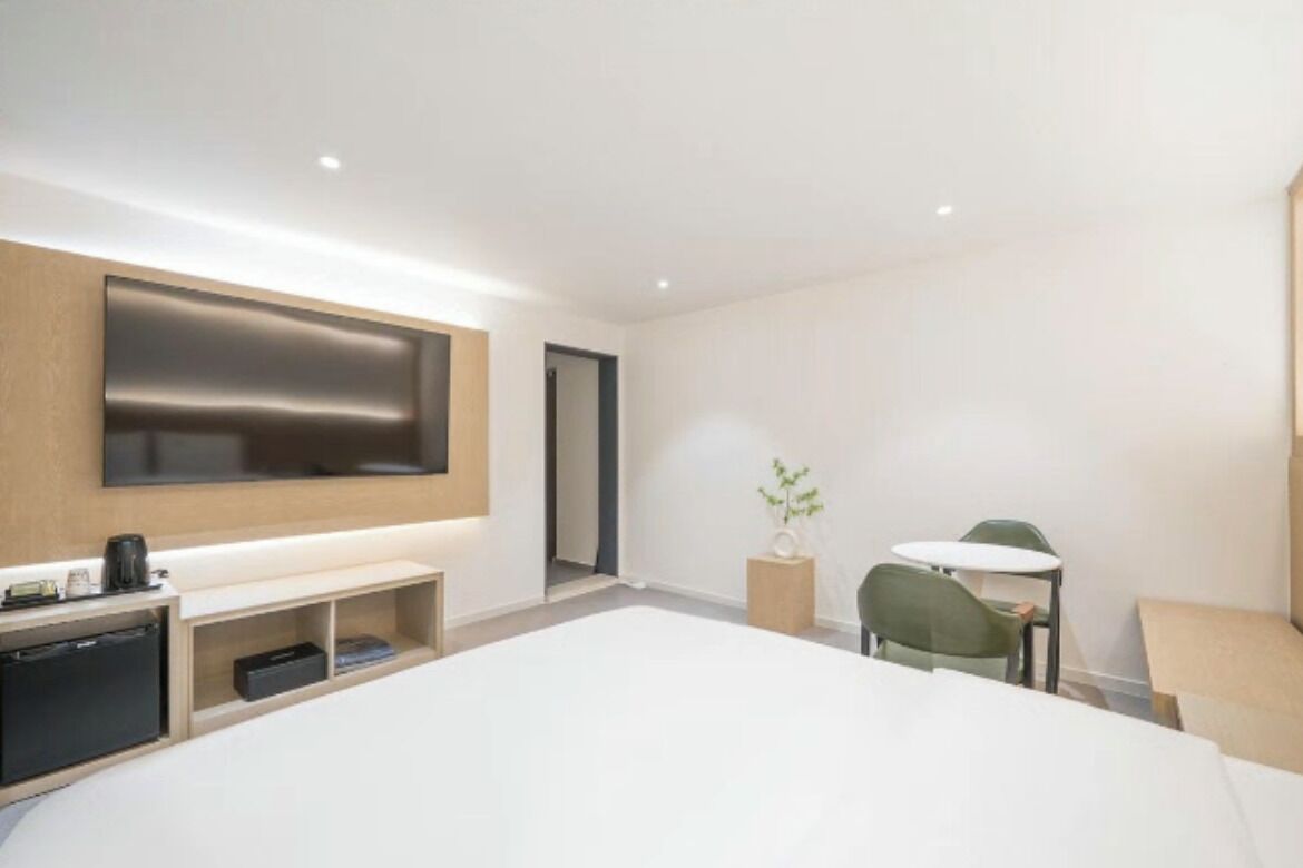 A modern hotel room with a large bed, a flat-screen TV mounted on the wall, a small desk with a chair, and a bedside table with a lamp.