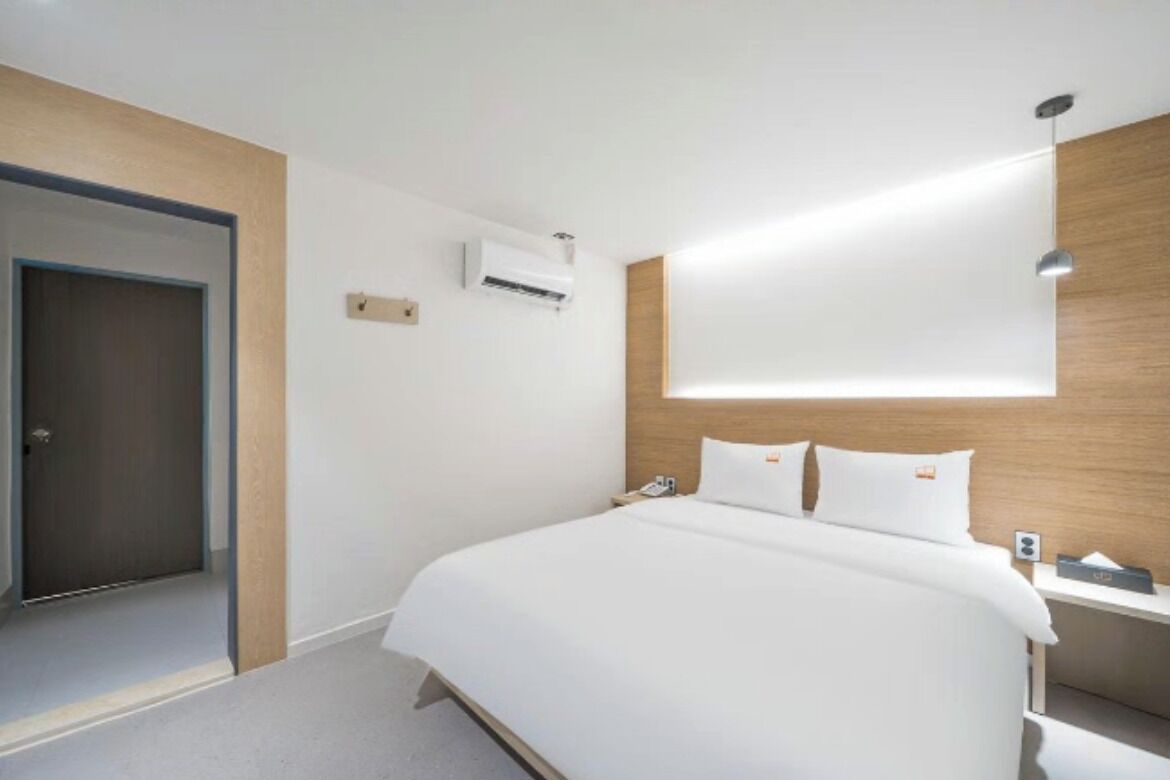 A hotel room with a bed, a bedside table, an air conditioning unit, and a door.