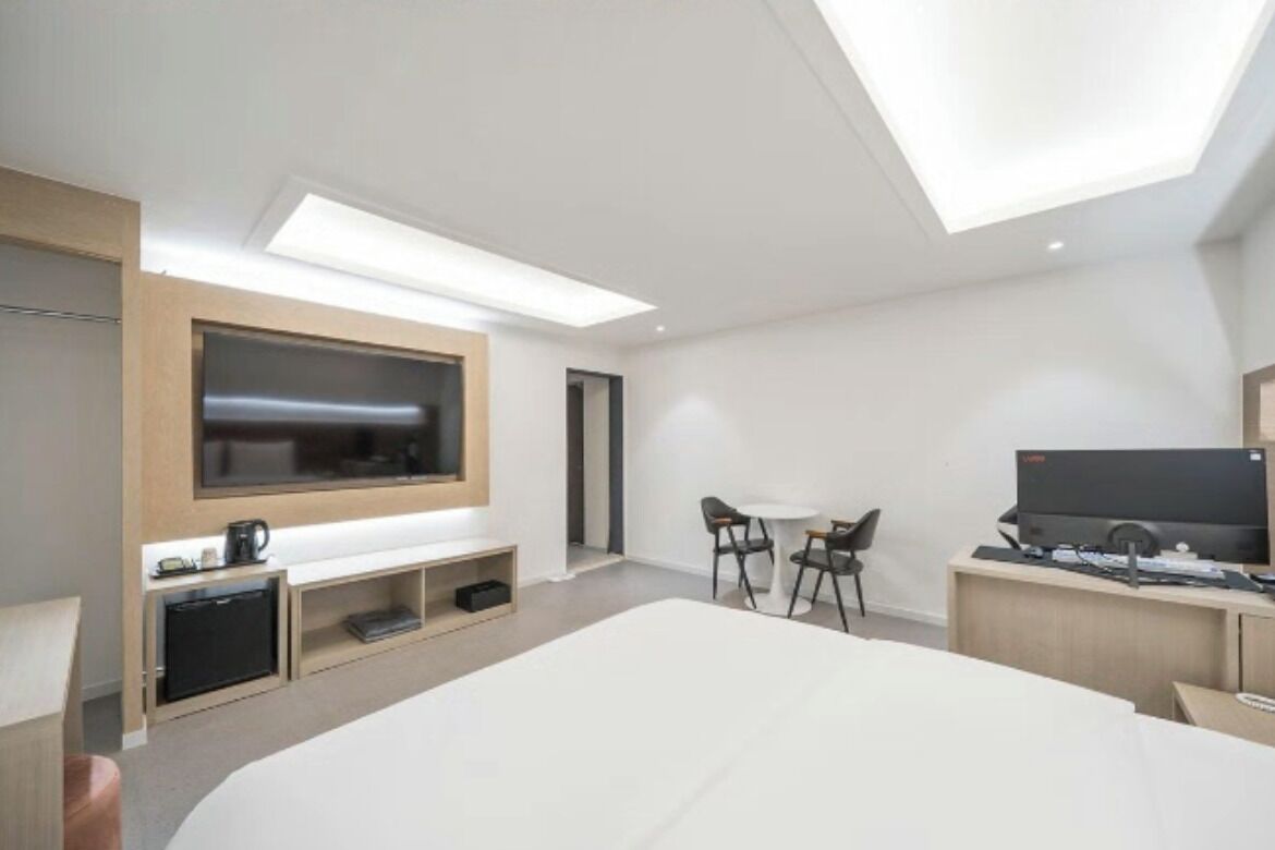 A modern hotel room with a large bed, a TV mounted on the wall, a desk with a computer, and a small seating area.