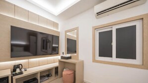 A modern hotel room with a large flat-screen TV, a small desk with a coffee maker, a pink ottoman, and a window with blinds. - DEUMEU HOTEL (Changwon)