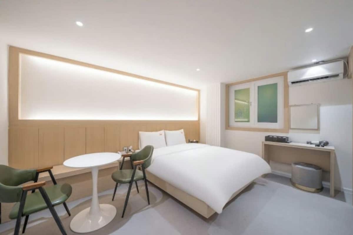 A hotel room with a bed, a round table, two chairs, a small desk, a trash bin, and a wall-mounted air conditioning unit.