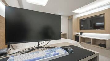 A modern hotel room with a large flat-screen TV, a desk with a keyboard and mouse, a bed, and a bedside table with a lamp.