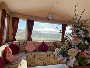 A cozy living area with a sofa, cushions, a clock, and a window with a view. - Cambrian Coast Holiday Park - Holiday Accommodation 14157 (Borth)