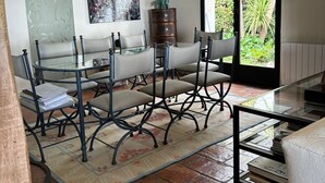 A dining area with a glass table, metal chairs, a patterned rug, a cityscape painting, and a window with a view of greenery. - Villa BOUGAINVILLEA, magnificent villa on the heights of Vence. (Vence)