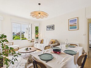 A dining area with a table set for two, a sofa, and a view of the garden. - 450m from the beach, suitable for 2 adults/2 child (Brignogan-Plage)