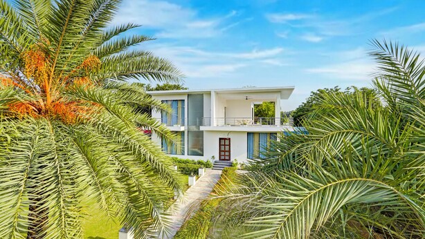 A modern two-story house with a central entrance, surrounded by lush palm trees and a clear sky. - Alaya Stays Tranquil Meadows 5BHK Farm (Taoru)