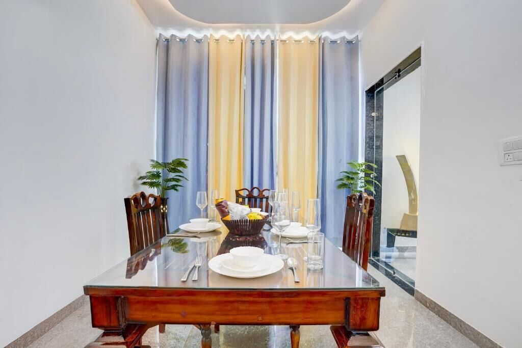 Una mesa de comedor con platos, vasos y una cesta de fruta, flanqueada por sillas, en una habitación con un gran ventanal y cortinas.