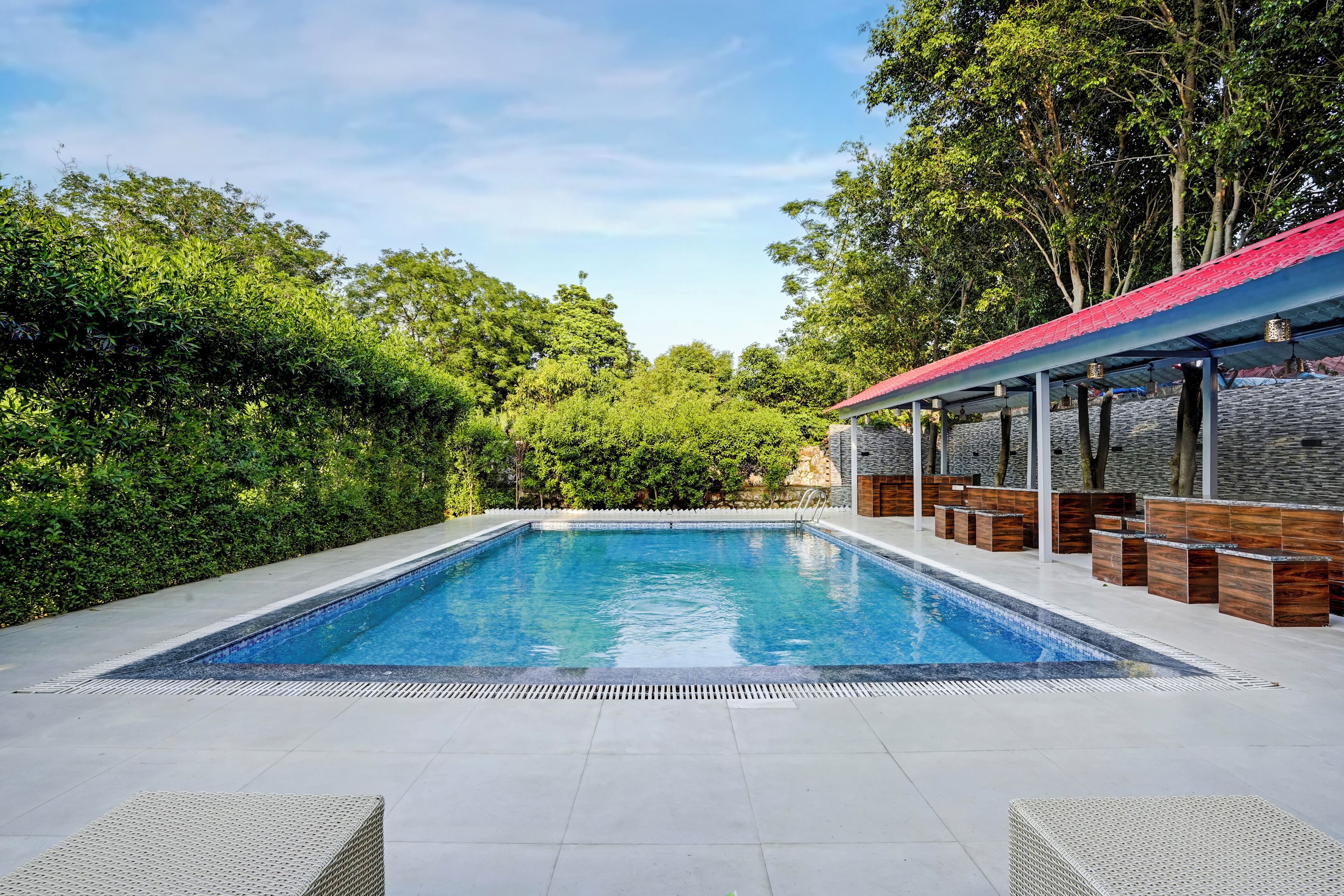 A rectangular swimming pool with a tiled deck, surrounded by lush greenery and a stone wall.