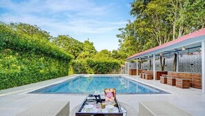 A poolside area with lounge chairs, a tray with drinks and snacks, and a stone wall with wooden seating. - Alaya Stays Tranquil Meadows 5BHK Farm (Taoru)