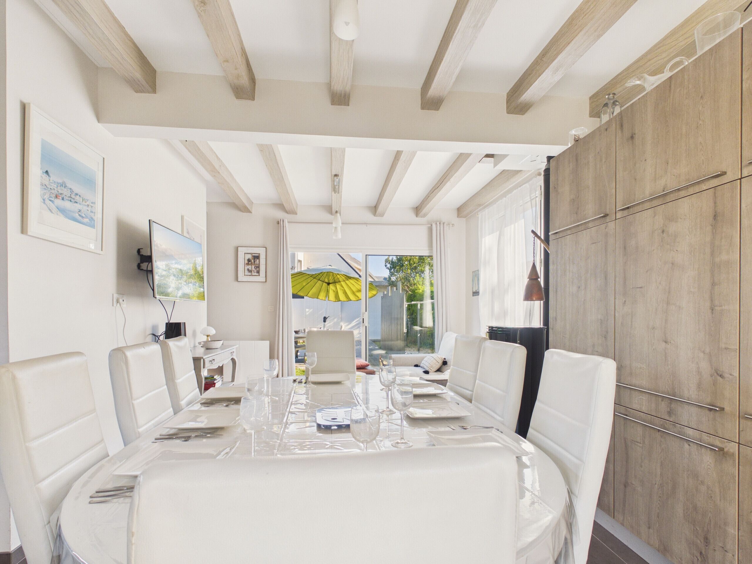A dining area with a table set for four, white chairs, a wooden ceiling, and a view through the glass door.