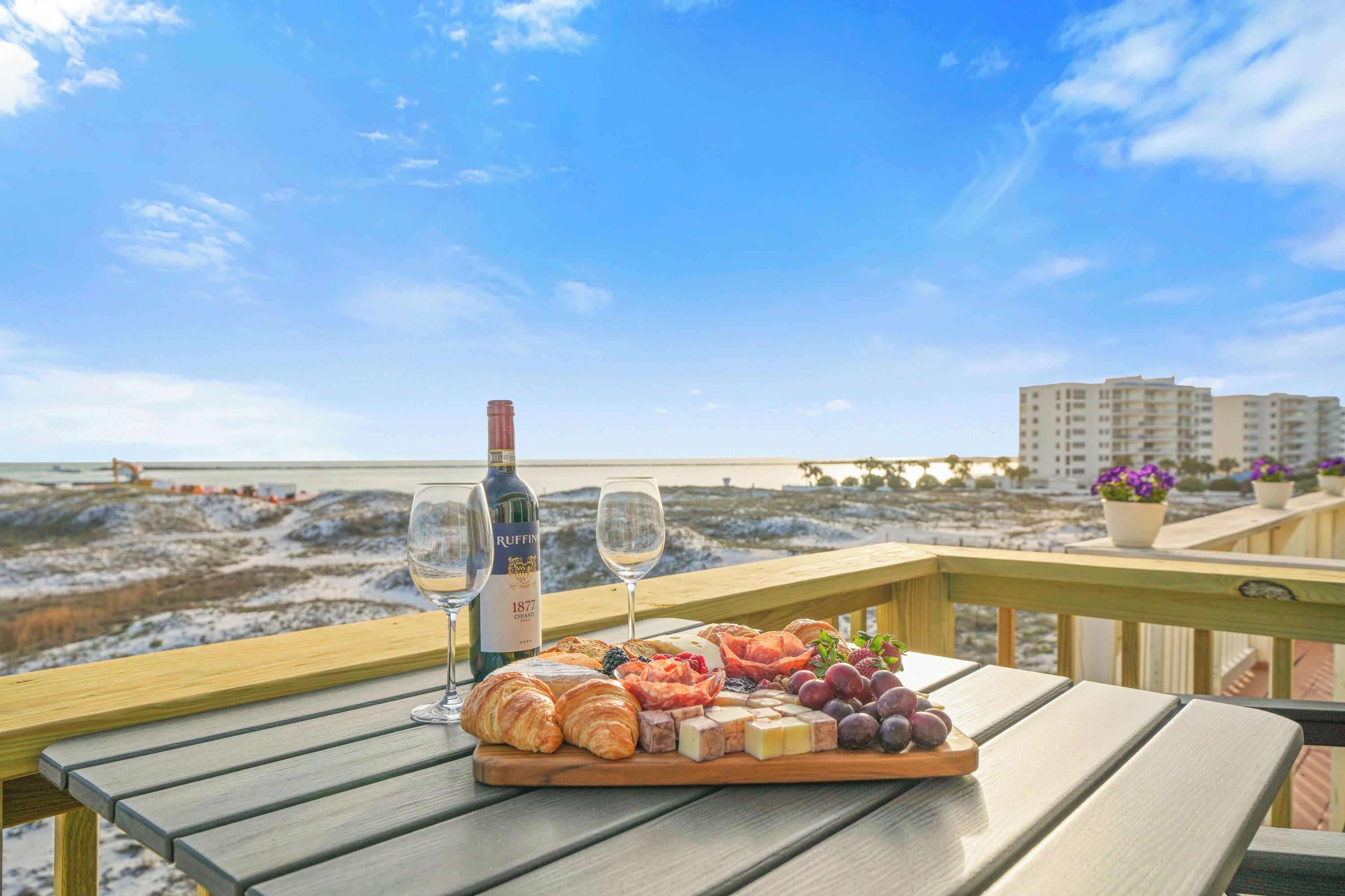 A wooden table with a charcuterie board, two wine glasses, and a bottle of wine on a balcony overlooking a beach.