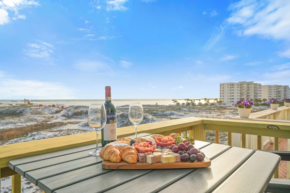 A wooden table with a charcuterie board, two wine glasses, and a bottle of wine on a balcony overlooking a beach.