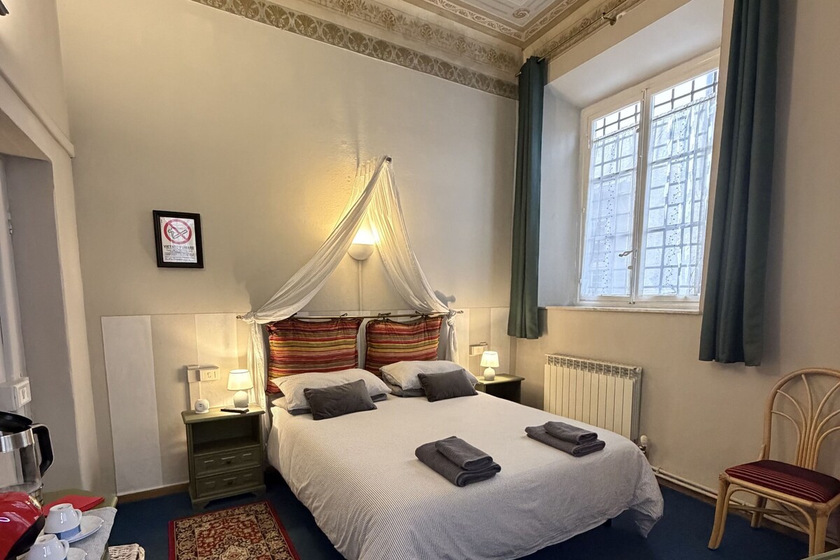 A neatly arranged hotel room with a large bed, bedside tables, a nightstand, a chair, a window with curtains, and a decorative ceiling.