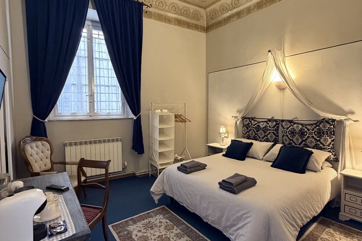 A bedroom with a bed, a desk, a chair, a radiator, and a window with curtains.