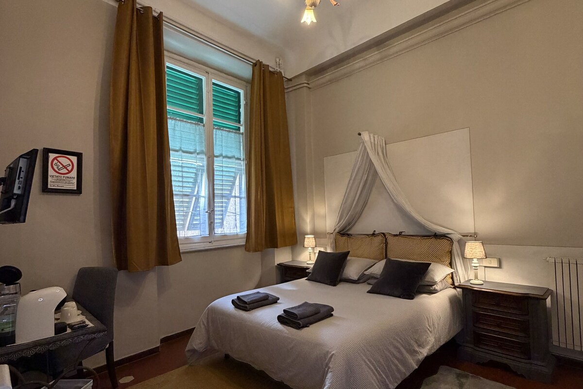 A bedroom with a bed, bedside tables, a desk, a chair, a window with curtains, a chandelier, and a radiator.