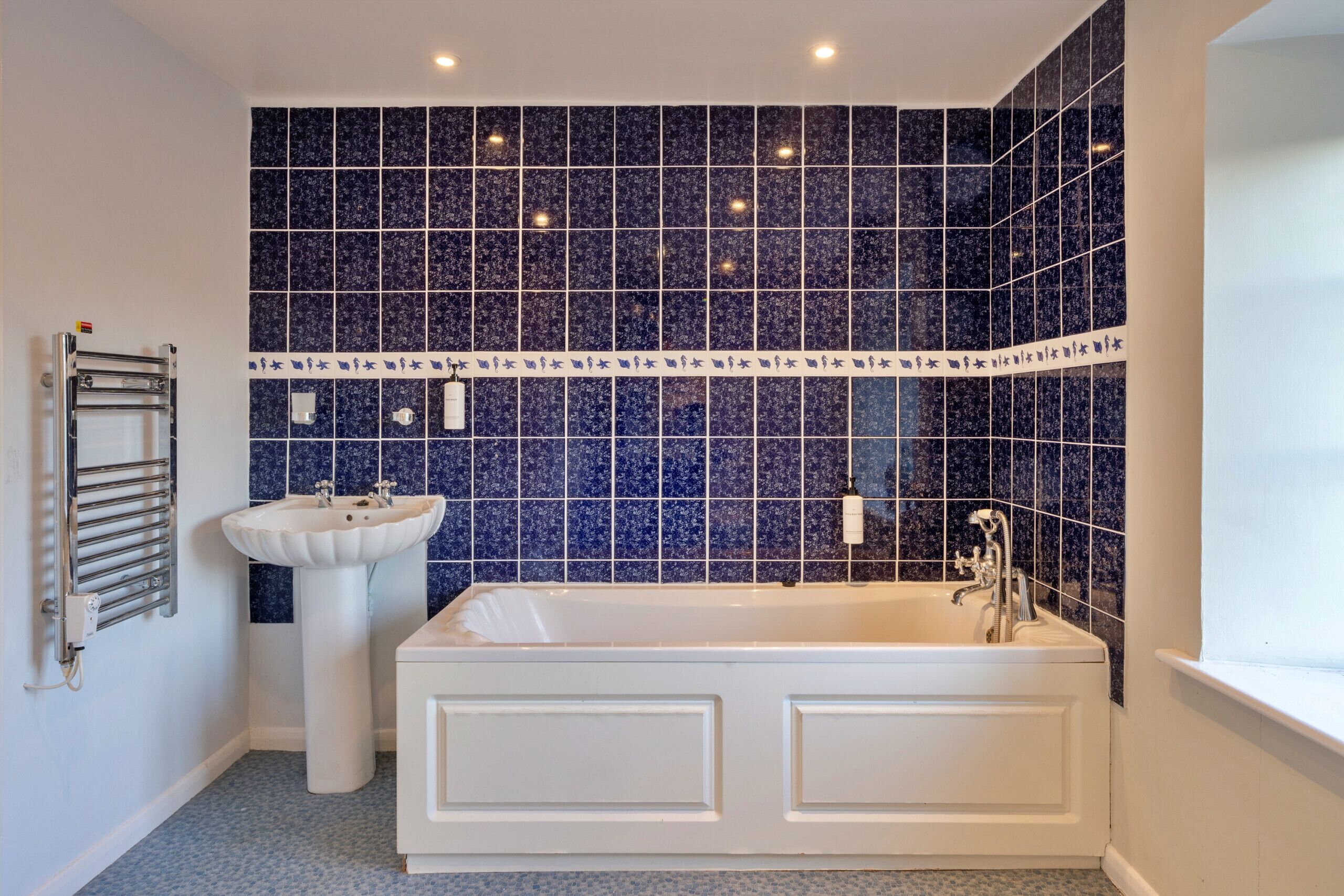 A bathroom with a white bathtub, a white pedestal sink, and a wall tiled with blue tiles.