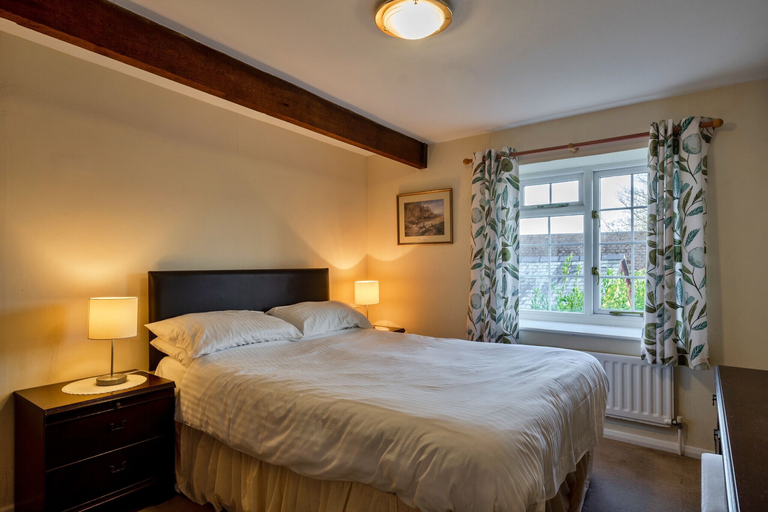 Une chambre à coucher avec un lit, des lampes de chevet, une fenêtre avec des rideaux, un radiateur et un tableau encadré au mur.