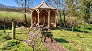 Ein hölzerner Gazebo mit Ziegeldach, umgeben von einem Kiesweg und einem Picknicktisch.