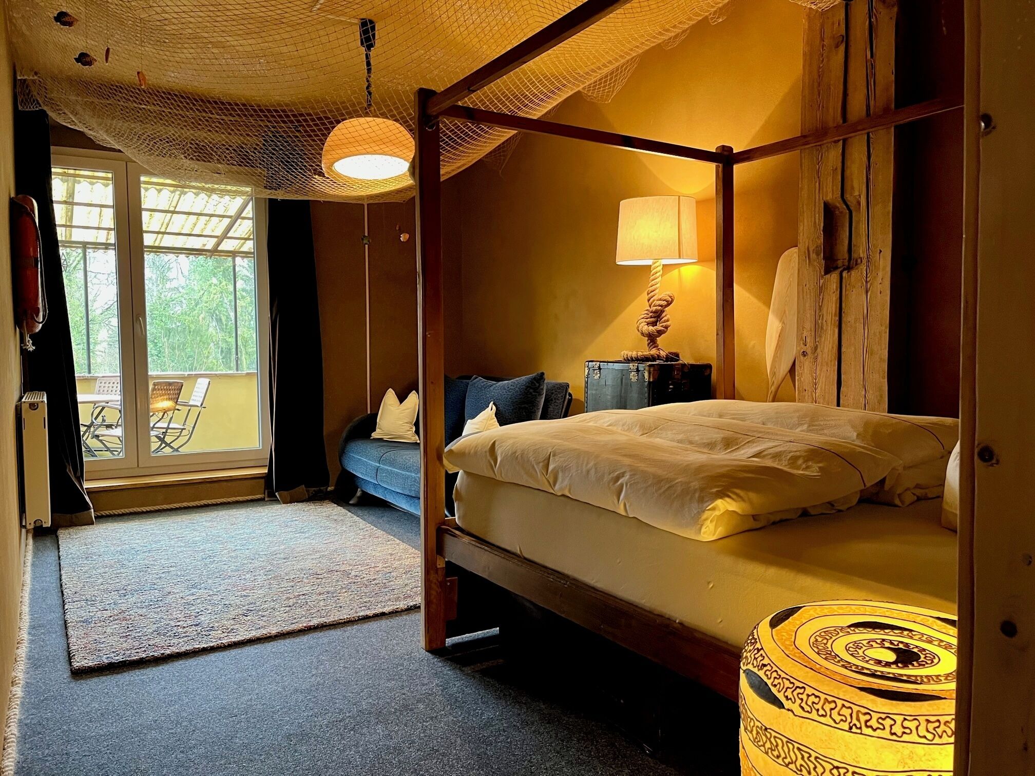 Un lit à baldaquin avec une lampe, une petite table et un banc dans une pièce donnant sur l’extérieur grâce à une porte vitrée.